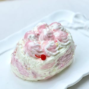 May include: A cake-shaped candle with white and pink frosting swirls, topped with a red cherry. The candle sits on a white, scalloped-edge plate. Sprinkles add a touch of sparkle. The candle is approximately 10 cm in length.