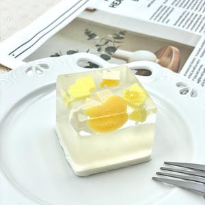 May include: A clear, cube-shaped dessert with yellow fruit pieces, including lemon slices and orange segments, sits on a white plate. The dessert is surrounded by a white base. A fork is visible on the right. The background includes a newspaper with the text "I HAVE YOU IN MY HEART I MISS YOU BY ERWHER".