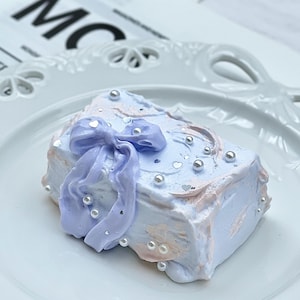 Puede incluir: Un pequeño objeto decorativo rectangular de color pastel que se asemeja a un pastel, adornado con un lazo lavanda y cuentas perladas. El artículo está sobre un plato blanco adornado. Las letras "MC" son visibles en el fondo.