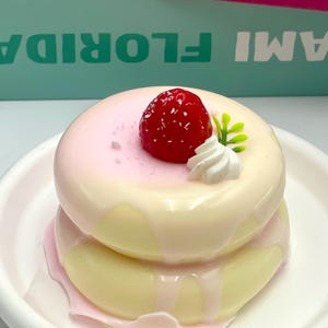 May include: A two-tiered dessert resembling a donut, with a light yellow base and a pink glaze. It's topped with a red strawberry, a dollop of white cream, and green leaves. The dessert sits on a white plate.
