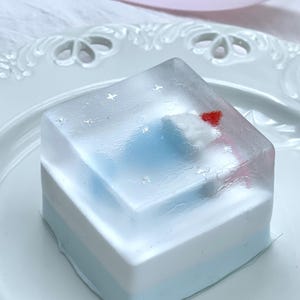 May include: A layered dessert with a clear, jelly-like top. The layers are blue, white, and a lighter blue. Tiny star-shaped sprinkles and a red heart decorate the top. The dessert is presented on a white, decorative plate.