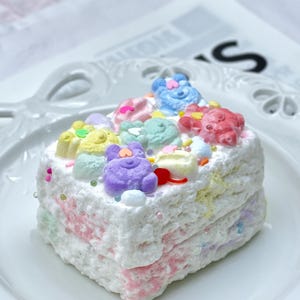 May include: A square cake with layers of pastel-coloured frosting, decorated with bear-shaped sweets and sprinkles. The cake sits on a white plate. The cake has pink, yellow and blue frosting.