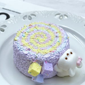 Puede incluir: Un pastel enrollado glaseado de colores pastel, con un diseño en espiral en tonos amarillos, morados y azules. El pastel está decorado con chispas y purpurina en forma de estrella. Un personaje blanco de dibujos animados con un lazo rosa está al lado del pastel.