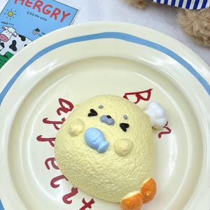 May include: A yellow seal-shaped ornament with a blue fish, black eyes, and orange flippers, on a cream plate with red lettering and a blue rim. A blue and white striped fabric is visible nearby. The plate has a diameter of approximately 20 cm.