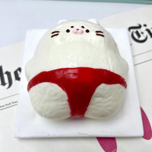 May include: A white, cat-shaped figurine with a red sumo wrestler's mawashi. The cat has black whiskers and eyes, and a pink nose. The figurine is on a white square base.