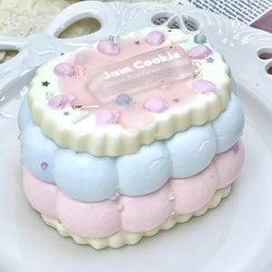 May include: A layered dessert resembling a cookie, with pastel pink, blue, and white layers. The top layer has the text "Jam Cookie" on a clear rectangular piece. Decorated with small pink accents, stars, and sprinkles, it sits on a white plate.