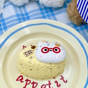 May include: A cream-coloured plate with a blue border features a small cake with a textured surface. The cake is decorated with a white cat figure wearing red glasses. The word "appetit" is written in red script.