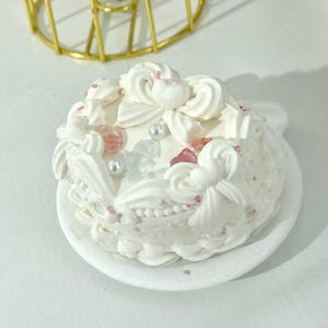 以下が含まれることがあります： 小さな白い装飾ケーキ。複雑なフロスティングの渦巻きとピンクと透明のアクセントがあり、お揃いの白いお皿に載っています。ケーキは小さな真珠のような装飾で飾られており、繊細でエレガントな外観を作り出しています。