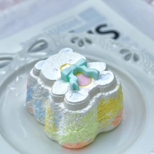 May include: A pastel-colored, bear-shaped bath bomb with a light blue bow on top. The bath bomb is white with pastel yellow, green, and orange accents. It sits on a white plate with a decorative edge.