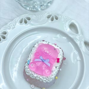 May include: A pink, rectangular dessert topped with a clear pink gummy bear and a blue bow. The dessert is decorated with white frosting and colourful sprinkles. It sits on a white plate.