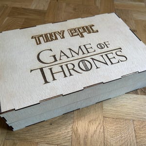 Może przedstawiać: Prostokątne pudełko z jasnego drewna z pokrywką zawierającą wygrawerowany laserowo napis Tiny Epic Game of Thrones. Pudełko posiada widoczne połączenia wczepowe wzdłuż krawędzi oraz gładką, płaską powierzchnię z naturalnym usłojeniem drewna.