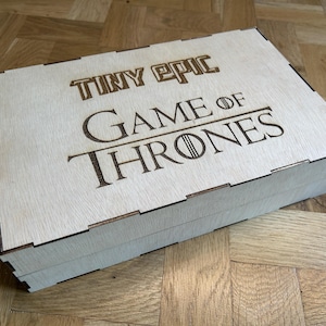 Może przedstawiać: Prostokątne pudełko z jasnego drewna z pokrywką zawierającą wygrawerowany laserowo napis Tiny Epic Game of Thrones. Pudełko posiada widoczne połączenia wczepowe wzdłuż krawędzi oraz gładką, płaską powierzchnię z naturalnym usłojeniem drewna.