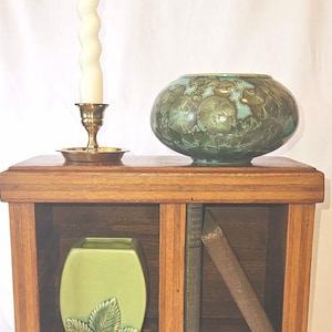 May include: A wooden cabinet with two shelves displays a green vase with leaf detailing, a decorative green and brown vase, a brass candlestick with a white spiral candle, and two vintage books. The cabinet is made of wood.