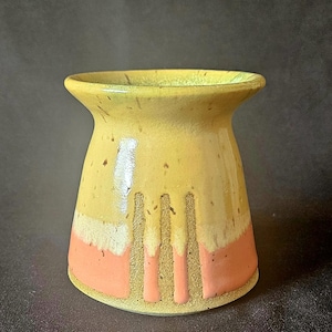 May include: A ceramic vessel with a flared rim, featuring a yellow upper section with dark speckles and a coral pink base. The design includes three vertical textured stripes that extend from the yellow glaze down into the coral pink area.
