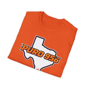 May include: An orange t-shirt featuring a white outline of the state of Texas with the words "PURO 956" in a stylized font. The shirt is made of a soft material and has a crew neck.