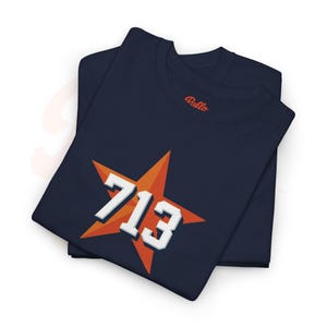 May include: Two navy blue t-shirts are folded and stacked. The top shirt features a large orange starburst design with the number "713" in white, 3D-style lettering. The brand name "Rallo" is embroidered in red on the collar.