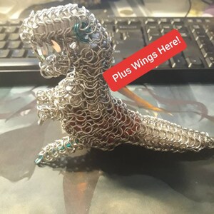 Peut inclure: Une sculpture de dragon en maille argentée avec des accents verts. Le dragon est en position debout, la tête inclinée vers le bas. La sculpture est sur un fond sombre avec le texte "Plus Wings Here!" écrit sur une étiquette rouge.