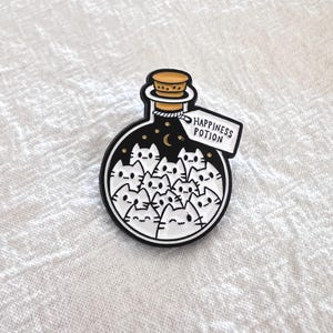 May include: A black and white enamel pin in the shape of a potion bottle. The bottle contains several white cat faces. A tag reads "Happiness Potion". The bottle has a cork top and a black background with stars and a crescent moon.