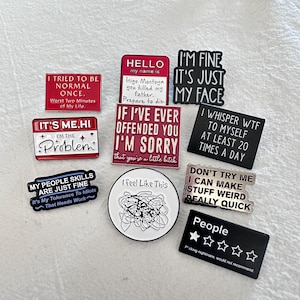 May include: A selection of enamel pins featuring witty and sarcastic sayings. The pins are in various shapes and colours, including red, black, and white. Text on the pins includes phrases such as "I tried to be normal once" and "I feel like this."