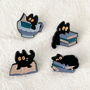 Może przedstawiać: Cztery czarne przypinki emaliowane z kotami. Jeden kot jest w filiżance do herbaty czytając książkę, inny siedzi na stosie książek, trzeci siedzi na otwartej książce, a ostatni śpi na stosie książek.