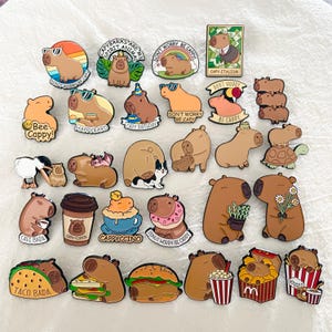 Pode incluir: Uma coleção de pins de esmalte com tema de capivara. Os pins apresentam capivaras com óculos de sol, segurando comida e com textos como "Don't Worry Be Cappy" e "Happy Bard".