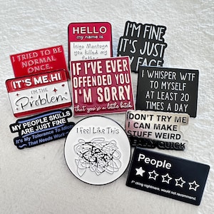 May include: A collection of novelty pins with humorous phrases and quotes. The pins are in various shapes and colors, including red, black, and white. Some pins feature text like "I'm Fine, It's Just My Face" and "I Feel Like This."