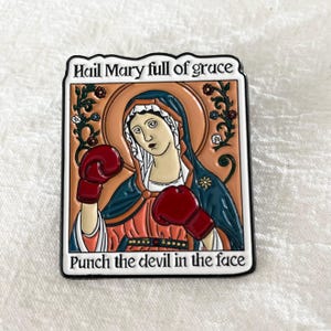 Może przedstawiać: Prostokątna przypinka przedstawiająca Maryję w czerwonych rękawicach bokserskich. Obrazek zawiera tekst "Hail Mary full of grace" na górze i "Punch the devil in the face" na dole. Grafika ma tematykę religijną.
