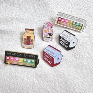 Peut inclure: Plusieurs épingles émaillées avec des motifs variés. L'une affiche "MY SOCIAL BATTERY" avec un indicateur de niveau de batterie. D'autres épingles présentent des bocaux avec des chats et des chiens, et des épingles en forme de glacière avec le texte "DON'T HATE ME BECAUSE I'M A Little Cooler."
