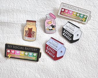 Peut inclure: Plusieurs épingles émaillées avec des motifs variés. L'une affiche "MY SOCIAL BATTERY" avec un indicateur de niveau de batterie. D'autres épingles présentent des bocaux avec des chats et des chiens, et des épingles en forme de glacière avec le texte "DON'T HATE ME BECAUSE I'M A Little Cooler."
