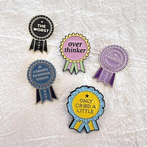 Op de afbeelding: Vijf kleurrijke emaille pins met lintdetails. De pins bevatten teksten als "The Worst", "Over Thinker", "I Talked to People Today!", "Conversation Awkward Participation" en "Only Cried a Little".