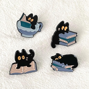 May include: Four black cat enamel pins. One cat is in a teacup reading a book, another is perched on a stack of books, a third is sitting on an open book, and the last is lying on a stack of books.
