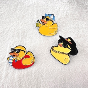 May include: Three yellow duck pins with black sunglasses. One duck wears a blue and white hat and holds a drink. Another wears a red shirt and a blue and white striped scarf. The third duck wears a black hat and a gold chain.