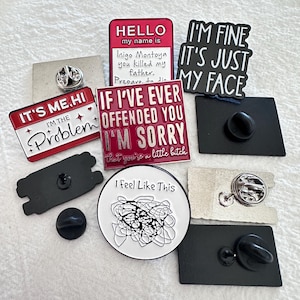 May include: A collection of enamel pins with various designs and text. The pins include phrases like "I'm fine, it's just my face," "It's me, hi, I'm the problem," and "If I've ever offended you, I'm sorry." Other pins feature abstract art and the text "Hello, my name is Inigo Montoya, you killed my father."