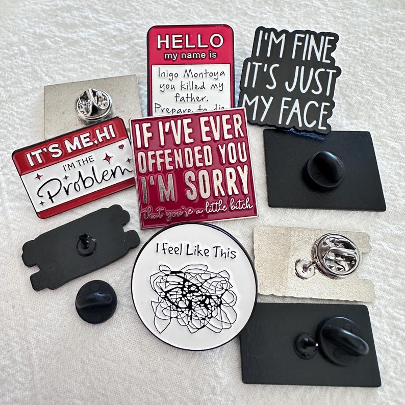 May include: A collection of enamel pins with various designs and text. The pins include phrases like "I'm fine, it's just my face," "It's me, hi, I'm the problem," and "If I've ever offended you, I'm sorry." Other pins feature abstract art and the text "Hello, my name is Inigo Montoya, you killed my father."