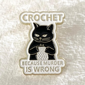 Peut inclure: Épingle noire représentant un chat tricotant avec du fil blanc. Le texte "CROCHET" est au-dessus et "BECAUSE MURDER IS WRONG" en dessous. Bordure blanche et dorée.