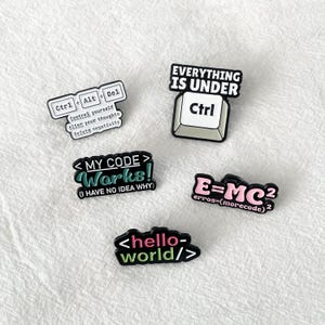 May include: Five enamel pins with IT-related designs. The pins feature keyboard keys such as Ctrl, Alt, and Del, and phrases like "My Code Works!" and "Hello World/". The colour scheme includes black, white, green, and pink.