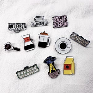 May include: A selection of coffee-themed enamel pins. The pins feature phrases like "But First Coffee" and "Need Coffee", alongside coffee-related imagery such as a coffee cup, a coffee maker, and a coffee bag. The pins are in various shapes and designs.