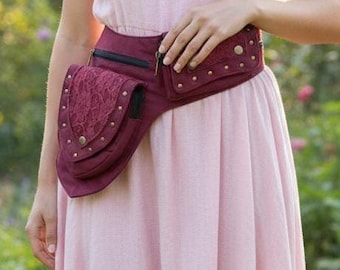 Puede incluir: Un cinturón de cintura burdeos con múltiples bolsillos y tachuelas decorativas. La bolsa presenta una solapa con un diseño en relieve y se usa sobre un vestido rosa claro. La bolsa está diseñada para usarse alrededor de la cintura.