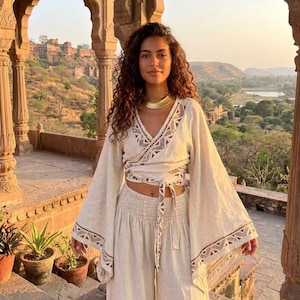 Puede incluir: Un top cruzado de color beige con mangas anchas y acampanadas y escote en V, con bordados geométricos de color marrón oscuro en los bordes y lazos en la cintura. Se combina con pantalones beige de talle alto y pernera ancha a juego.