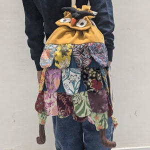 Puede incluir: Una mochila artesanal con forma de búho. La bolsa presenta un cuerpo amarillo mostaza, una falda de retazos con estampados florales y patas de tela marrón. La cara del búho tiene ojos blancos grandes, un pico negro y una cabeza negra.