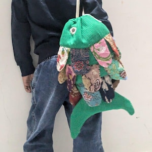 Può includere: Una borsa a tracolla verde a forma di pesce con un cinturino bianco. Il corpo della borsa è in tessuto verde e le pinne sono in tessuto fantasia colorato. La borsa è indossata a spalla.