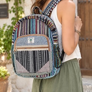 Può includere: Uno zaino in tessuto intrecciato multicolore con strisce verticali nei toni del blu, nero, bianco e bordeaux. Include una tasca frontale con cerniera e una toppa che mostra il testo HIMALAYAN RSH HEMP e l'icona di una foglia.