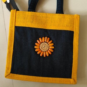 May include: A square tote bag with a yellow jute exterior and a navy blue front panel. The bag features a sunflower design made of wooden beads and a wooden button center. The handles are navy blue, and the bag has a zipper closure.