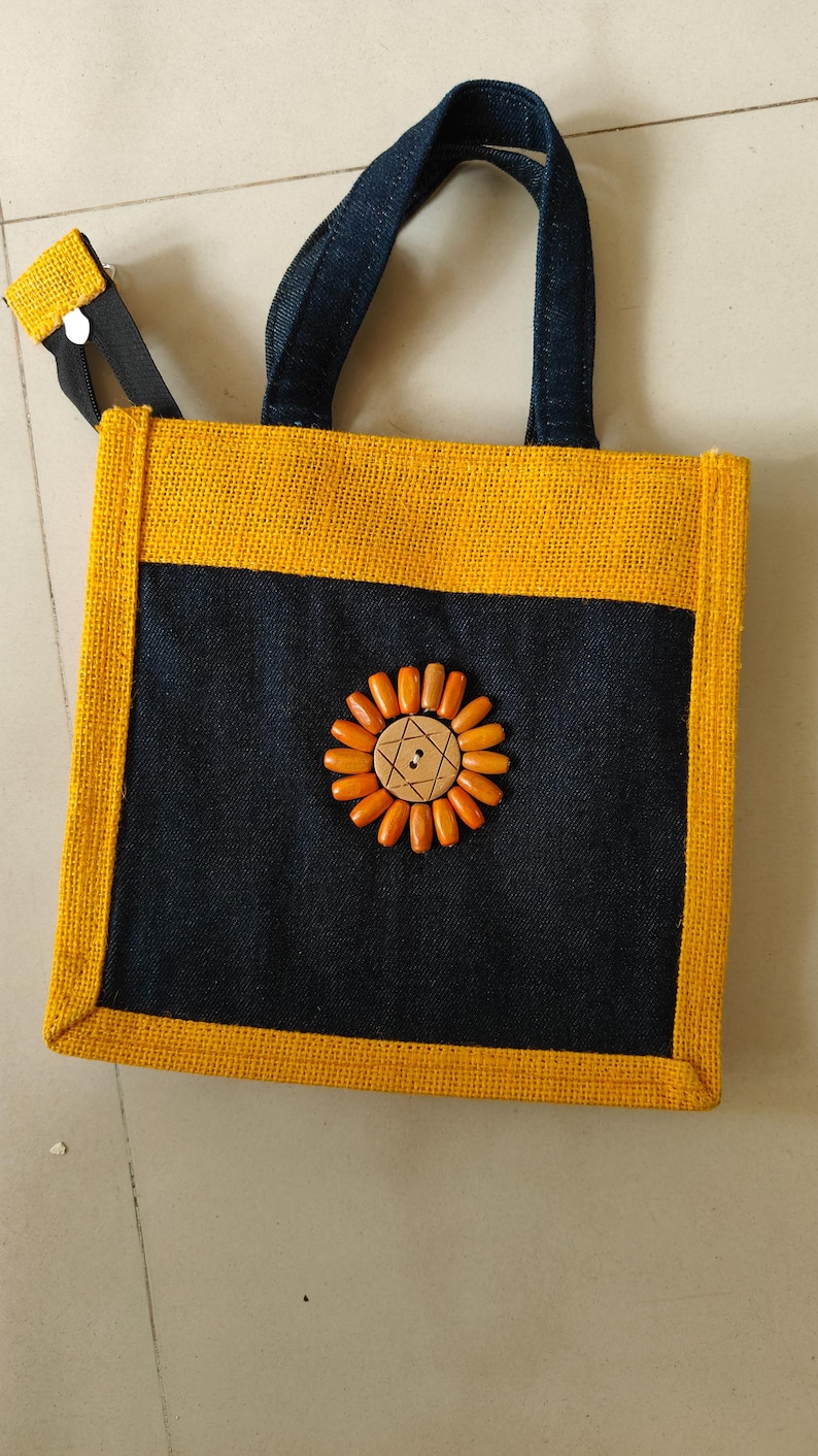 May include: A square tote bag with a yellow jute exterior and a navy blue front panel. The bag features a sunflower design made of wooden beads and a wooden button center. The handles are navy blue, and the bag has a zipper closure.
