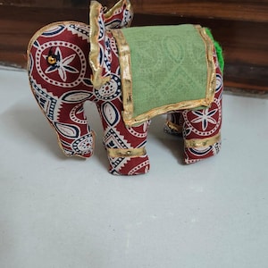 May include: A decorative stuffed elephant figurine. The elephant is mainly red with a white and navy blue floral pattern. It has a green saddle and gold trim. The elephant's ears are also trimmed with gold.