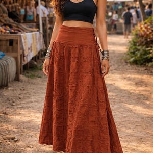 May include: A floor-length, rust-colored skirt with a textured, grid-like pattern and a side-tie waist detail. The skirt is paired with a black sleeveless crop top, a silver-toned statement necklace, and stacked silver-toned bracelets.