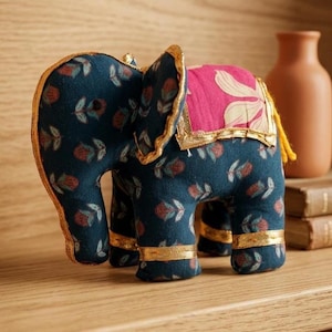 May include: A fabric elephant ornament in navy blue with a floral design and gold edging. It features a pink and white floral saddle. The ornament is displayed on a wooden surface.