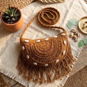 May include: A crescent-shaped mustard yellow crochet crossbody bag featuring a long shoulder strap and a fringe trim along the bottom edge. The front of the bag is decorated with several white cowrie shells arranged in a curved pattern.