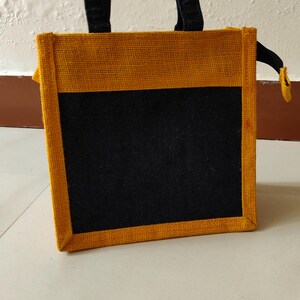 May include: A rectangular tote bag with a black front panel and a yellow-brown border and trim. The bag has black handles and a small yellow zipper pull. The bag is made of a woven material.