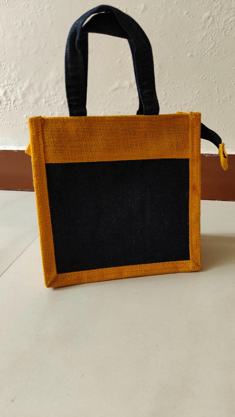 May include: A rectangular tote bag with a black front panel and a yellow-brown border and trim. The bag has black handles and a small yellow zipper pull. The bag is made of a woven material.